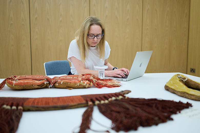 A blonde lady sits at a table using a magnifying glass to look closely at a textile object. Also on the table is a silver laptop.