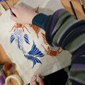 Young hand painting blue and orange Koi fish onto a tote bag