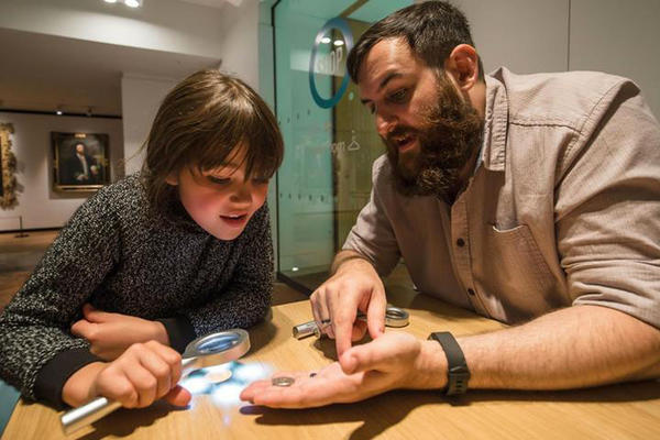 A young, interested child holds a magnifying glass and looks at a coin held in the palm of a man's hand. 