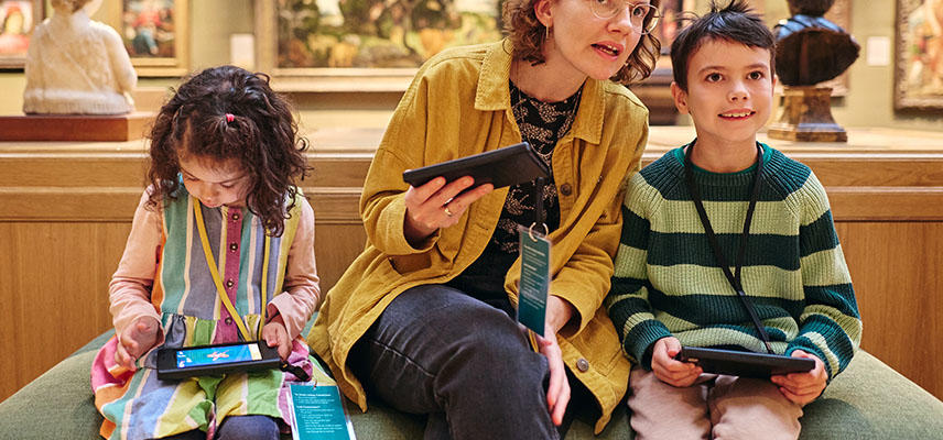 Two children and an adult sit on a green bench in a gallery space. Each hold tablets. 