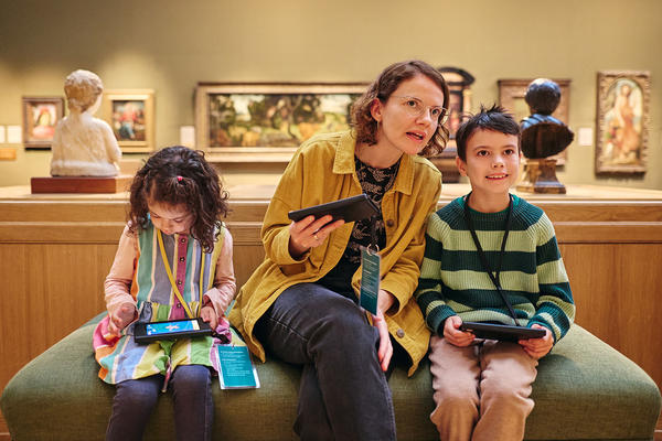 Two children and an adult sit on a green bench in a gallery space. Each hold tablets. 